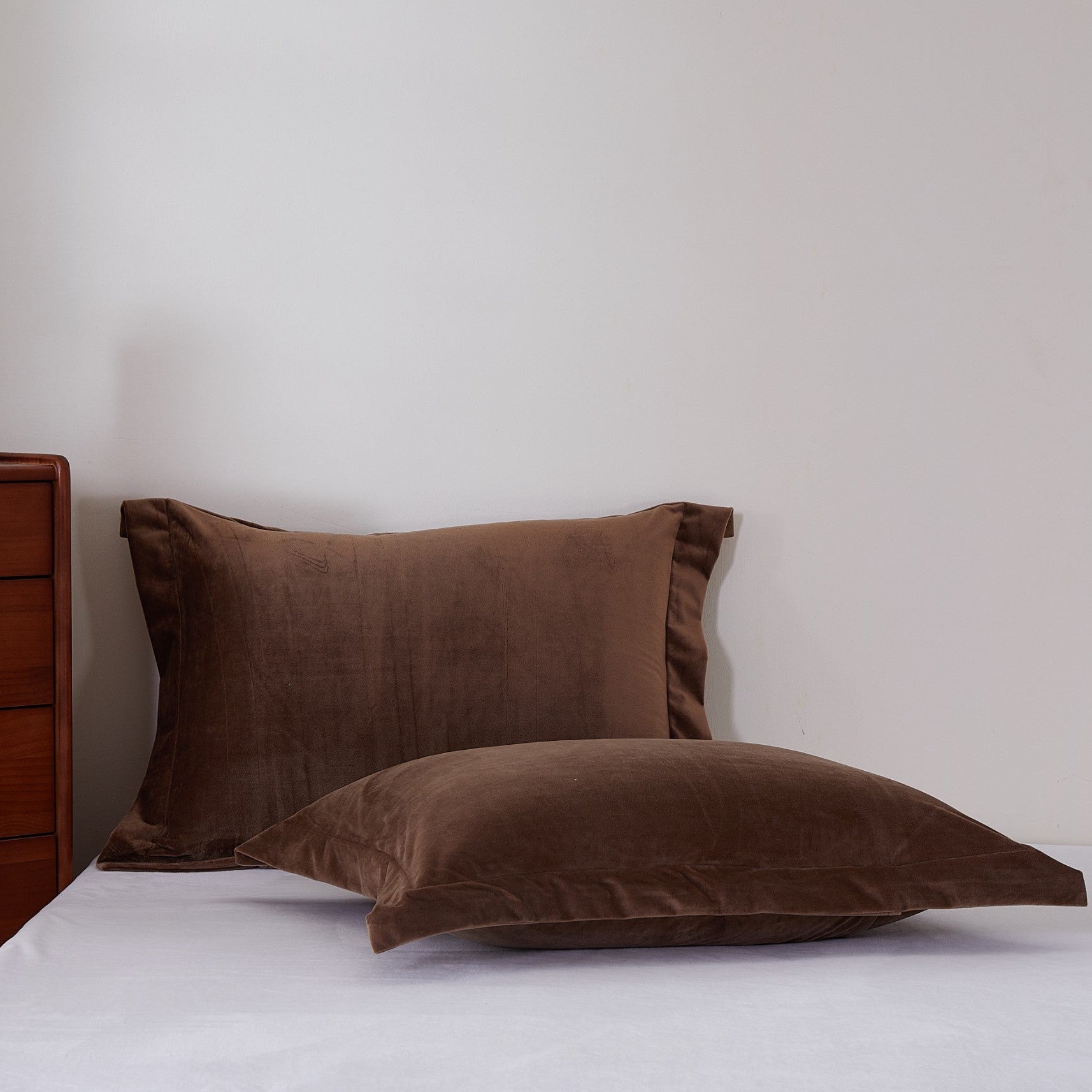 A set of brown pillowcases in different sizes, neatly stacked and placed on a bed next to a wooden nightstand with a framed picture on top.