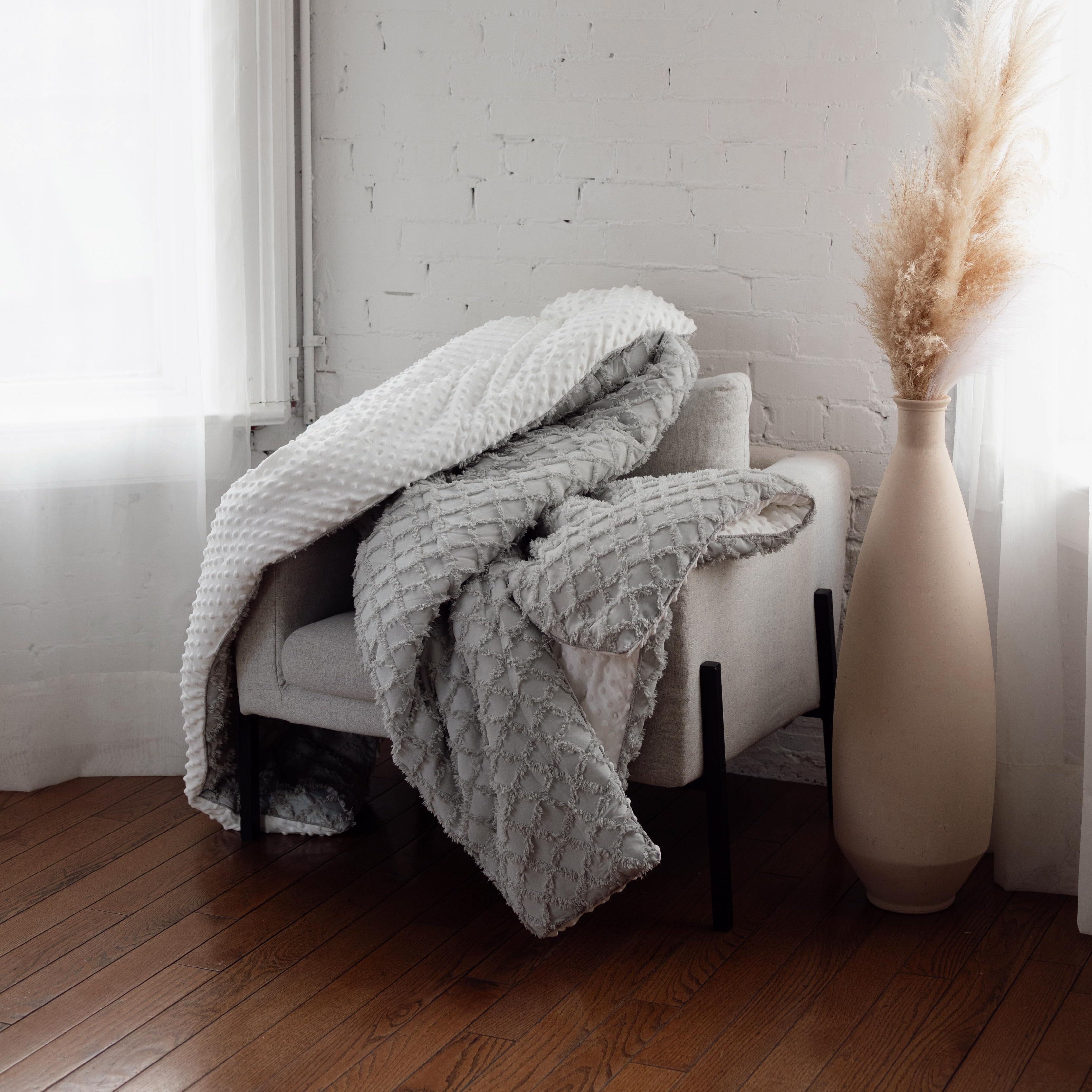 Light gray textured blanket draped over a chair next to a tall beige vase against a white brick wall.