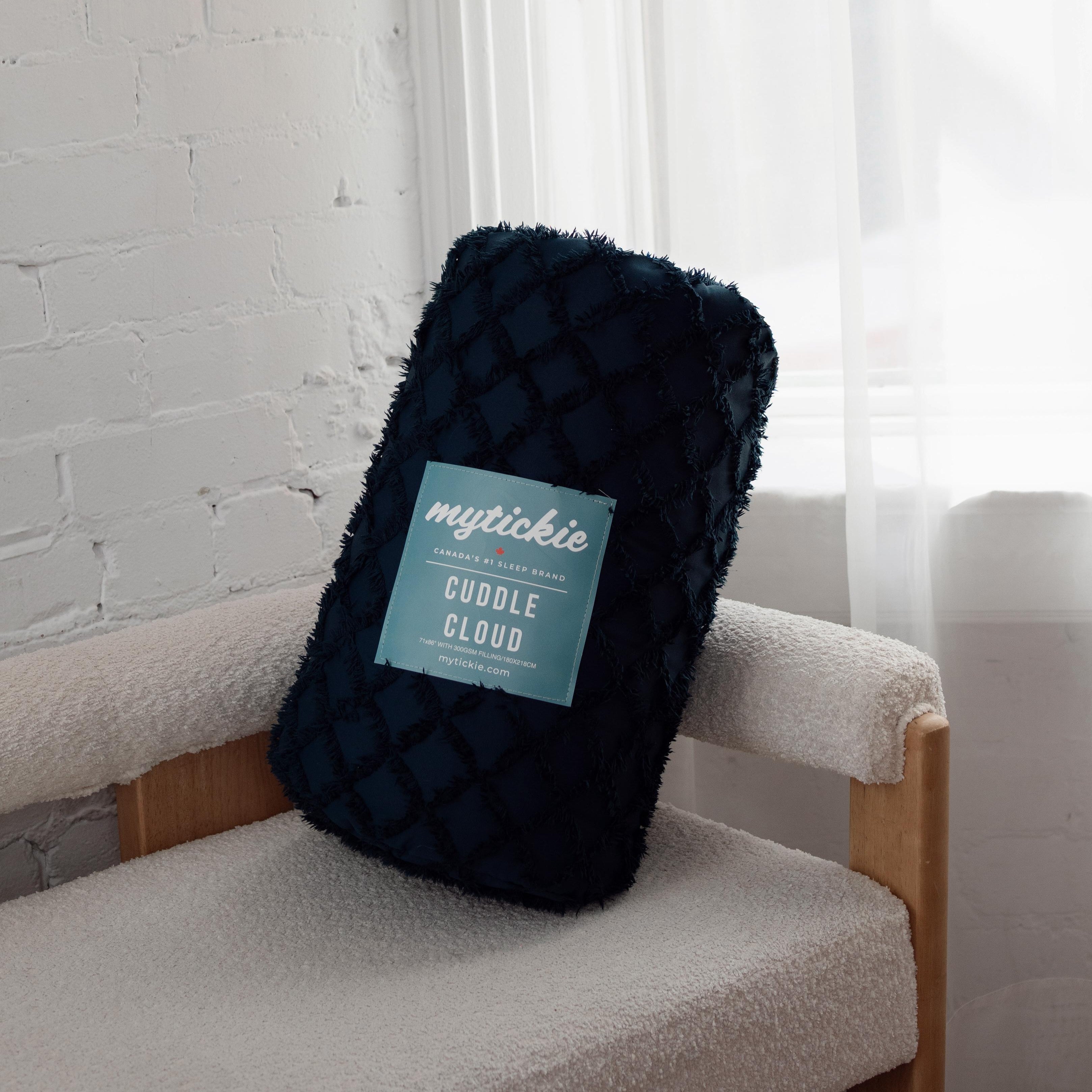 Dark blue throw blanket with a label on a chair against a white curtain and a brick wall.