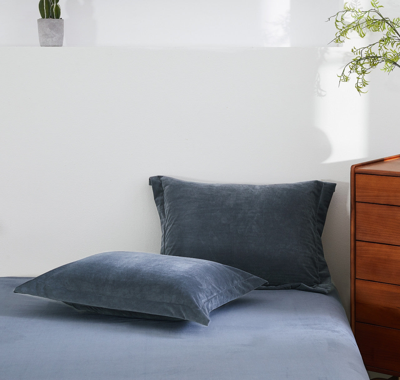 Gray pillows on a bed with a wooden dresser and plants in the background