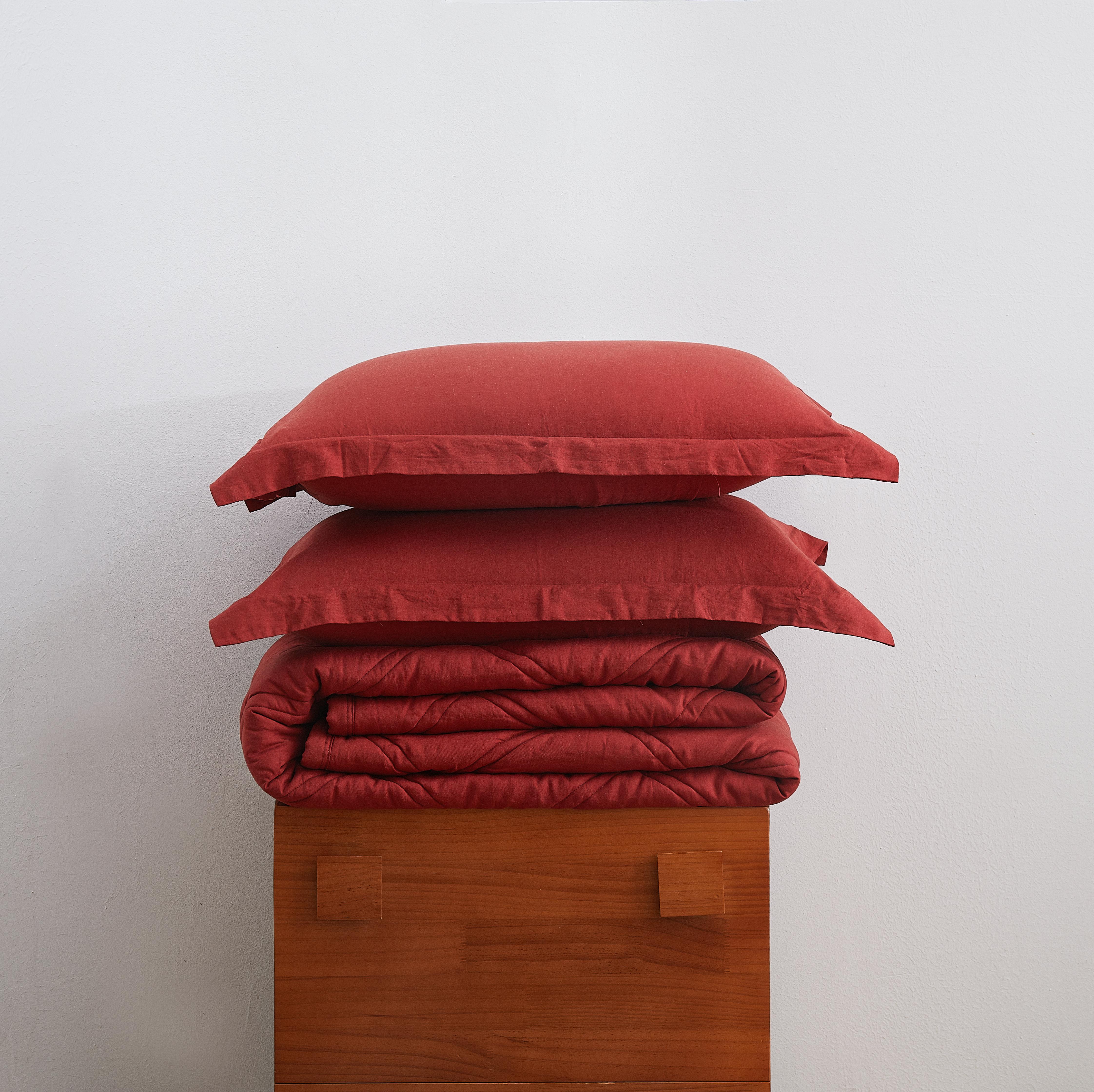 Red bedding set with two pillows and a folded blanket on a wooden stand against a white background