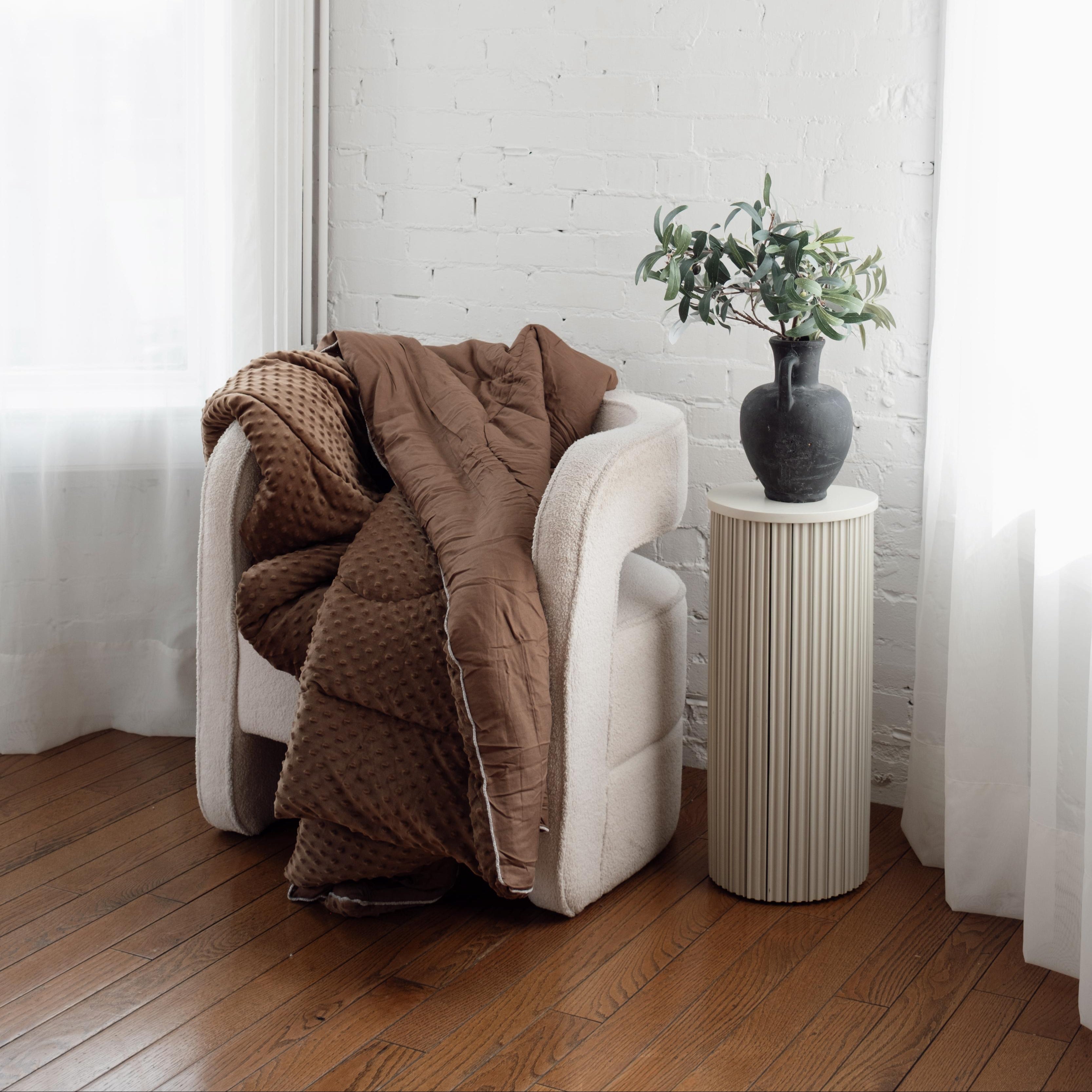 White armchair with a brown blanket and a small table with a plant against a white brick wall.
