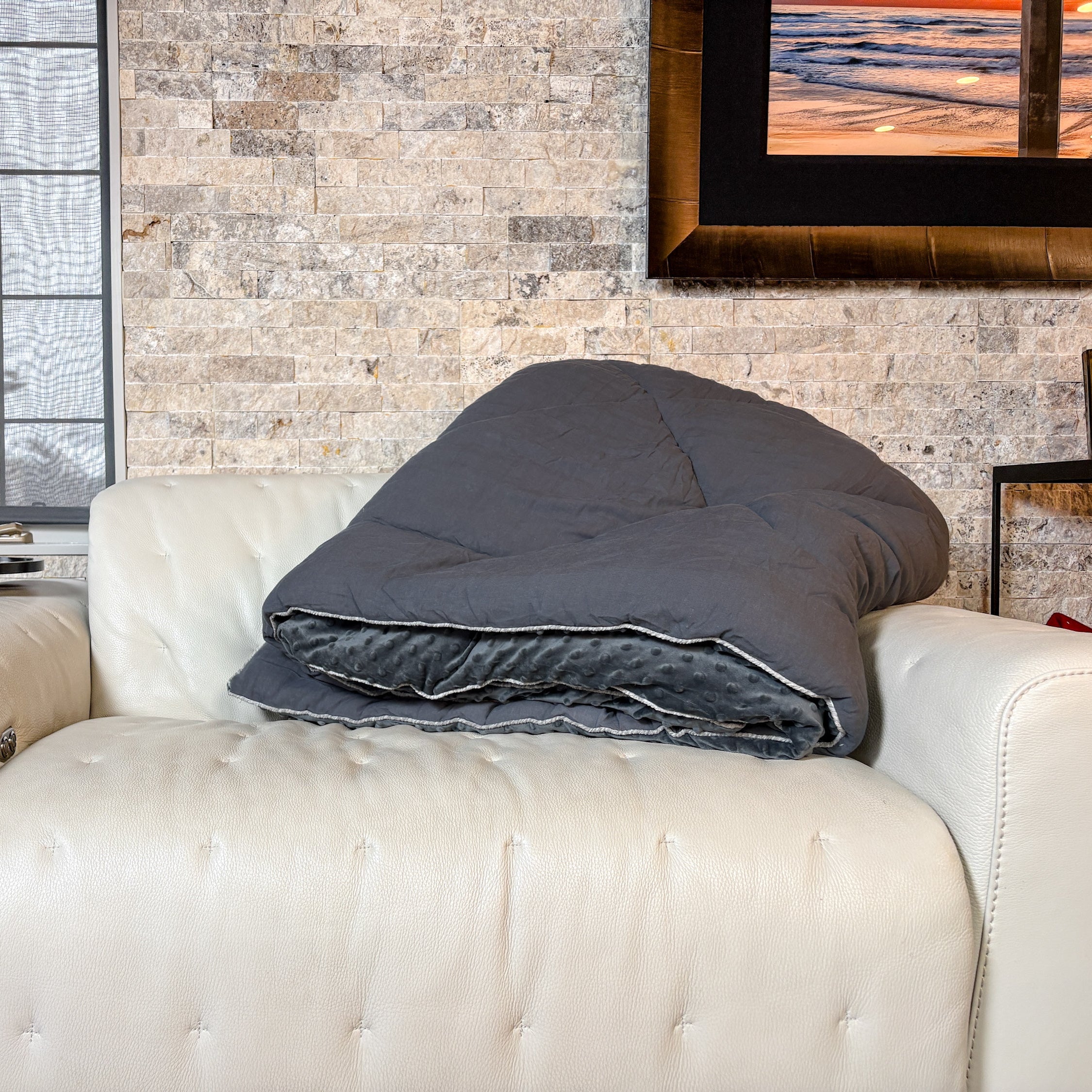 Gray comforter on a white sofa against a stone wall with a window and framed picture.