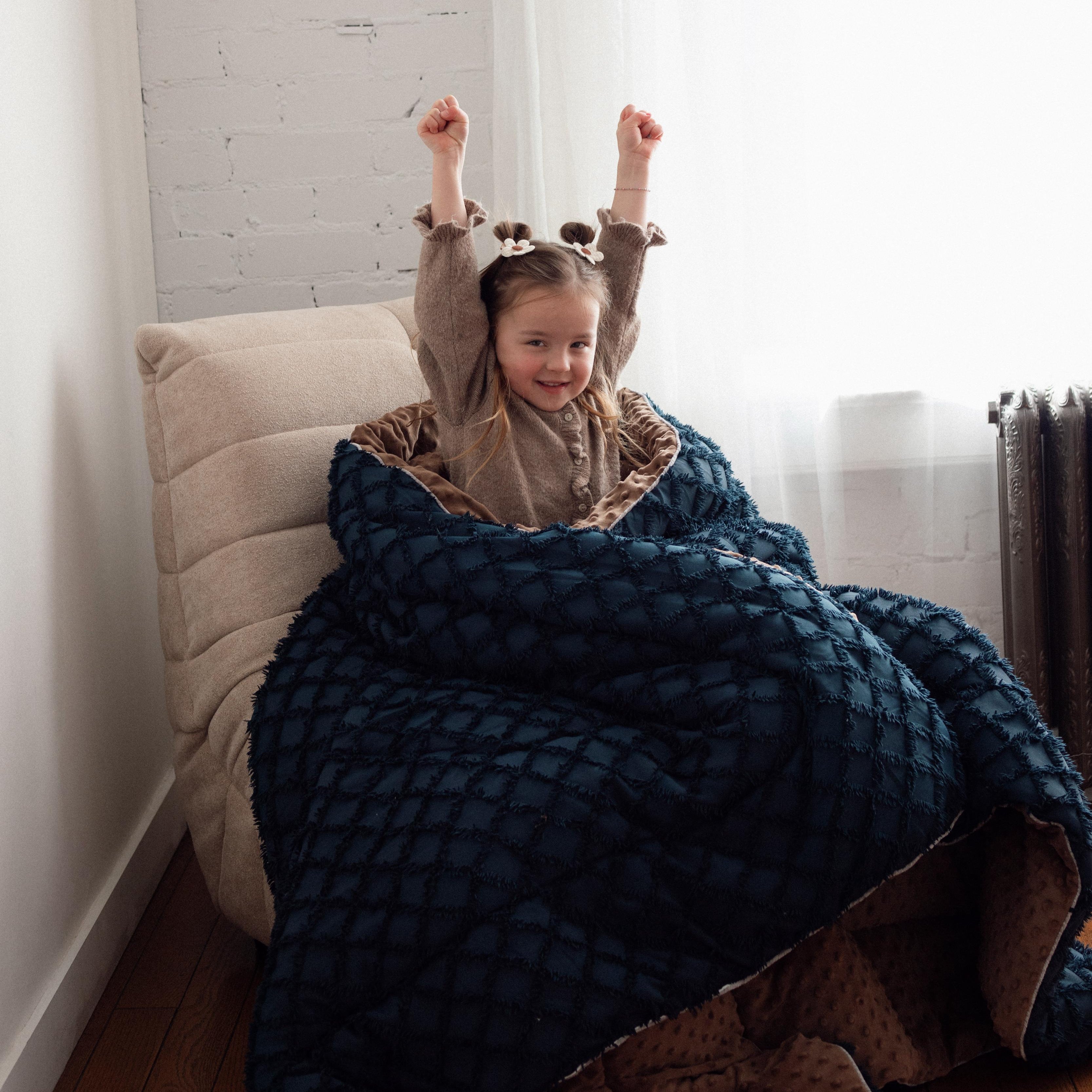 Child wrapped in a dark blue blanket sitting on a chair with arms raised in a room.