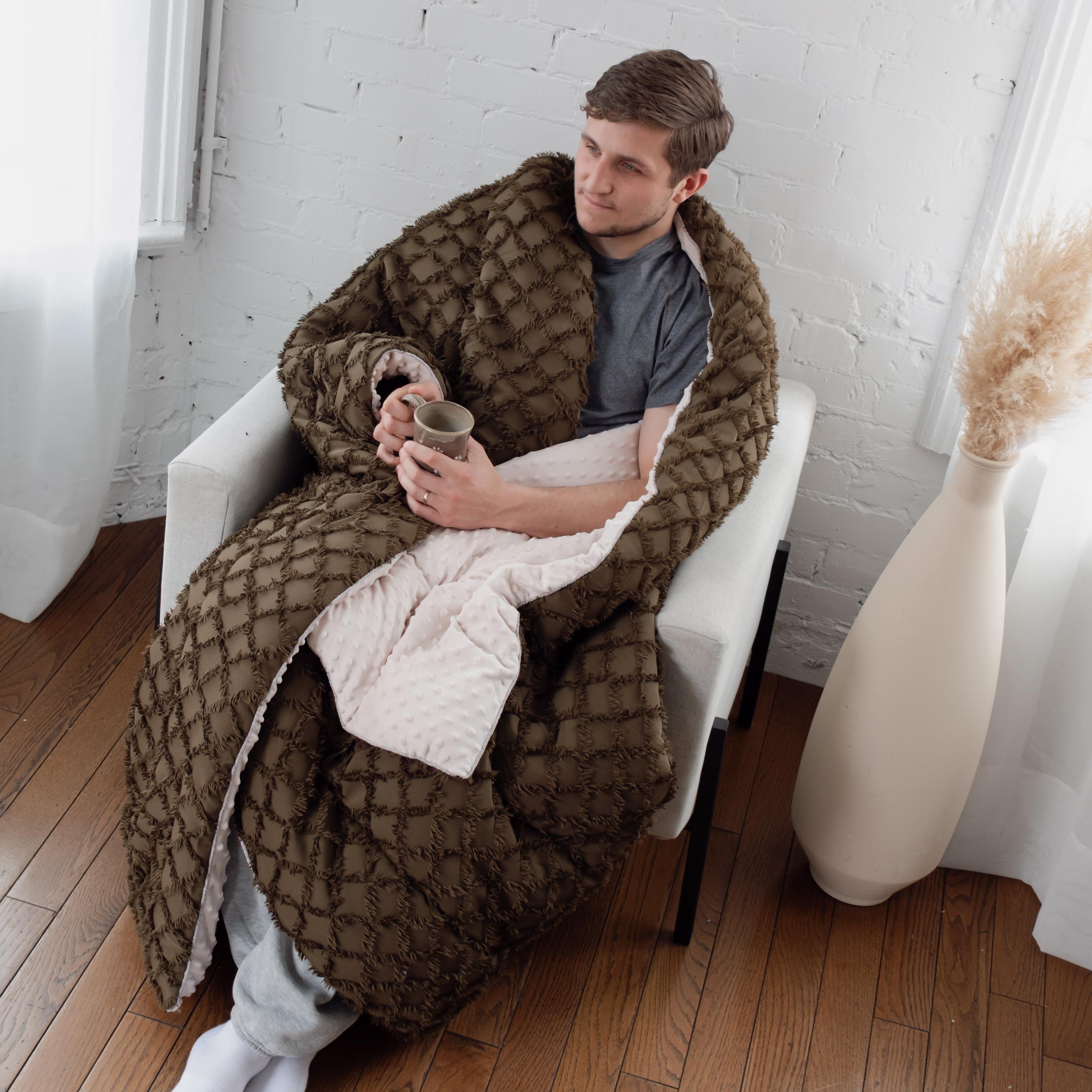 Person sitting on a chair wrapped in a large Olive throw blanket in a room with white walls and wooden floor.